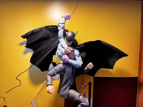 A recreation of the Detective Comics #27 cover using action figures, showing Batman swinging from the sky in a city holding a flailing criminal